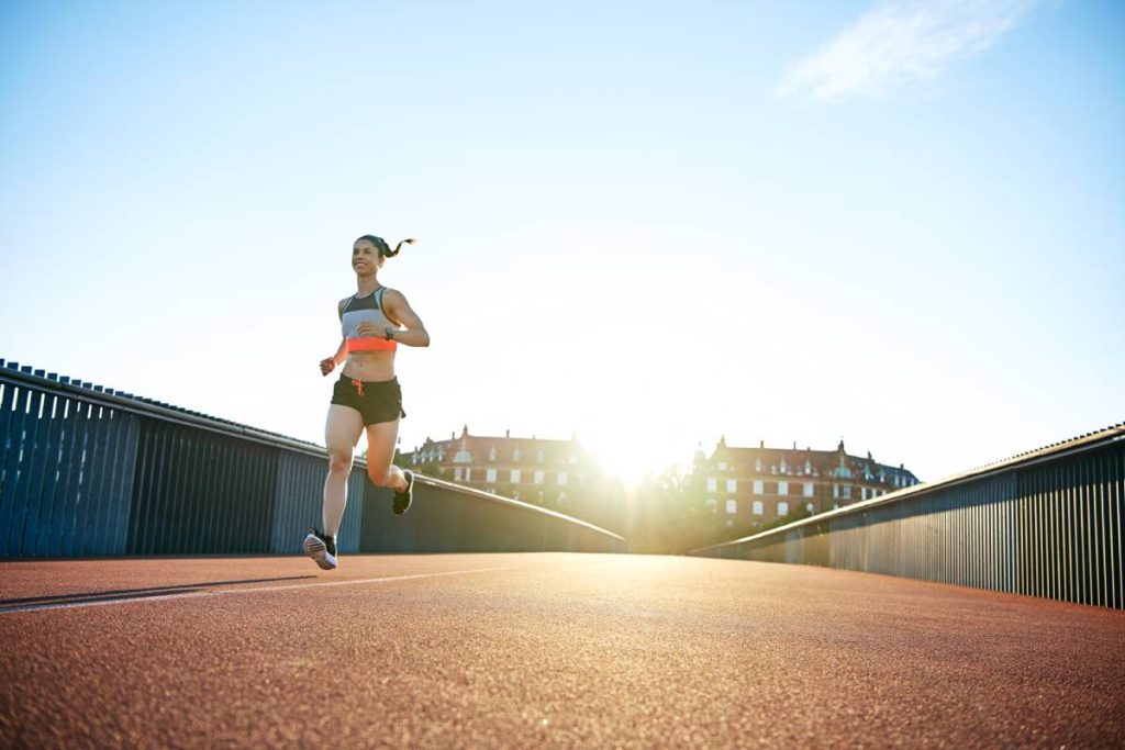 Low angle view of woman running down concrete and iron railing bridge with bright sunlight reaching over urban theme background