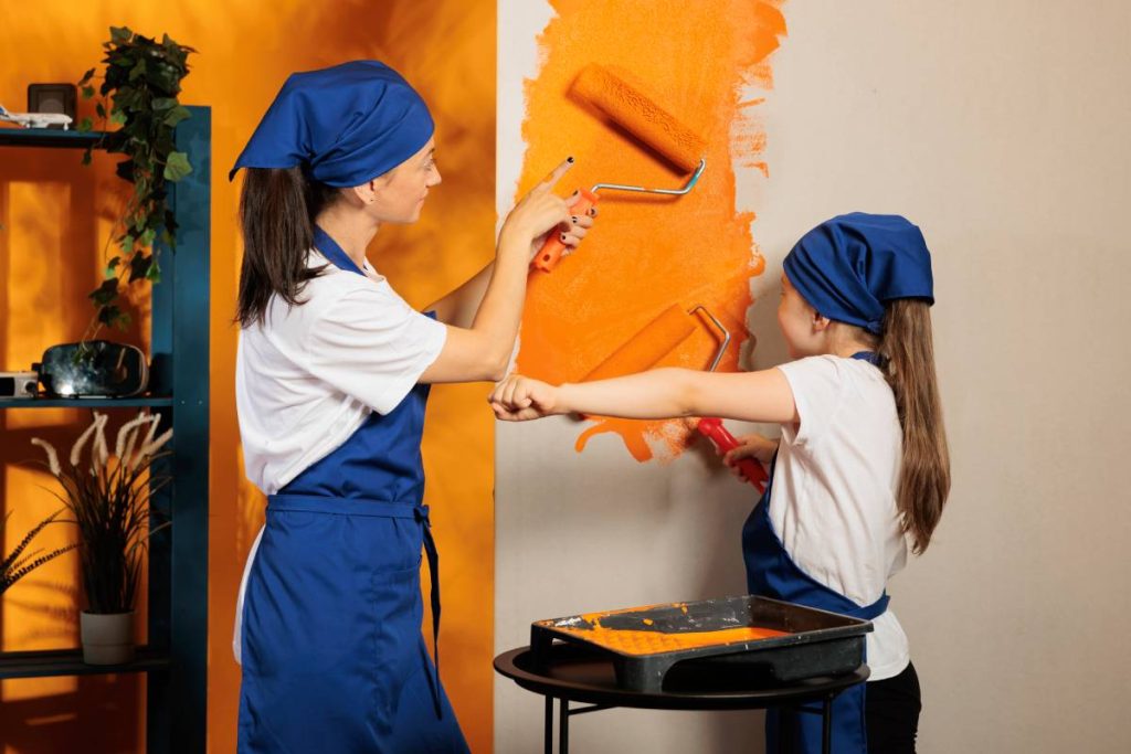 Mother and girl painting house walls with orange color paint, using renovating tools and paintbrush or roller to decorate apartment. Housework renovation with equipment, diy decoration.