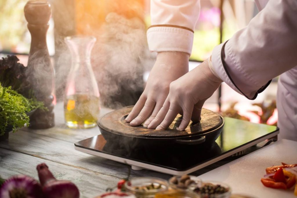 Wooden board covers frying pan. Hands of chef in uniform. It's a secret dish. Surprise for gourmets.