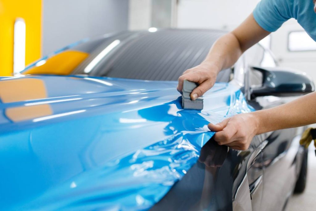 Car wrapping, mechanic with squeegee installs protective vinyl foil or film on vehicle hood. Worker makes auto detailing. Automobile paint protection coating, professional tuning