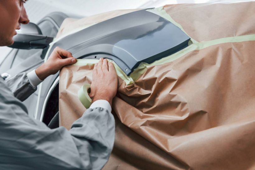 Covering car by brown cloth. Caucasian automobile repairman in uniform works in garage.