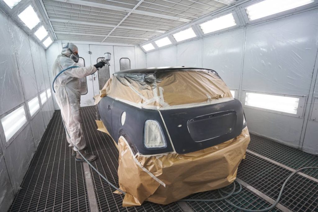 Car painter in white protective clothes standing in car painting booth, holding spray gun and painting car