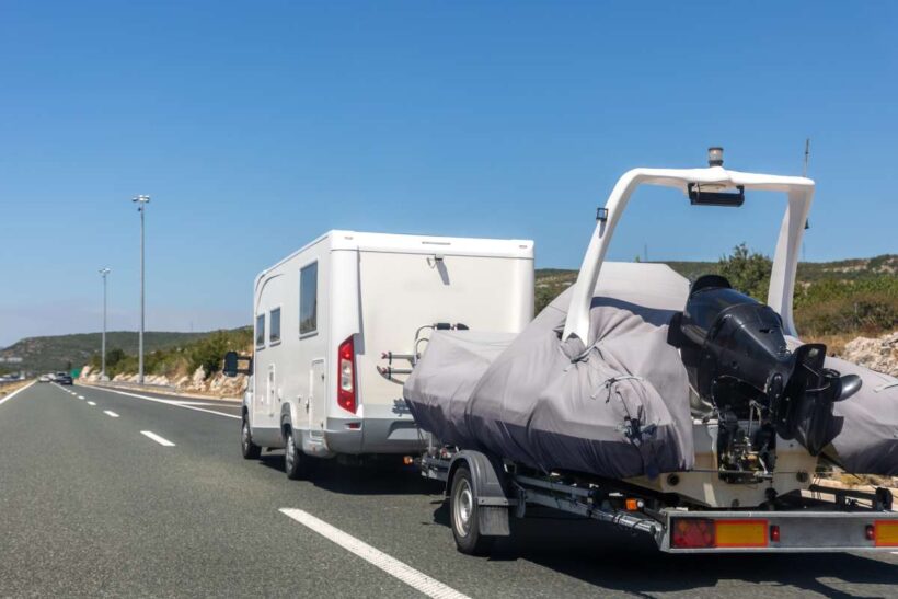 Motorhome towing covered boat trailer drives along empty highway clear blue sky surrounded by hills. Bright summer day creates sense freedom travel adventure mobile lifestyle on scenic route.
